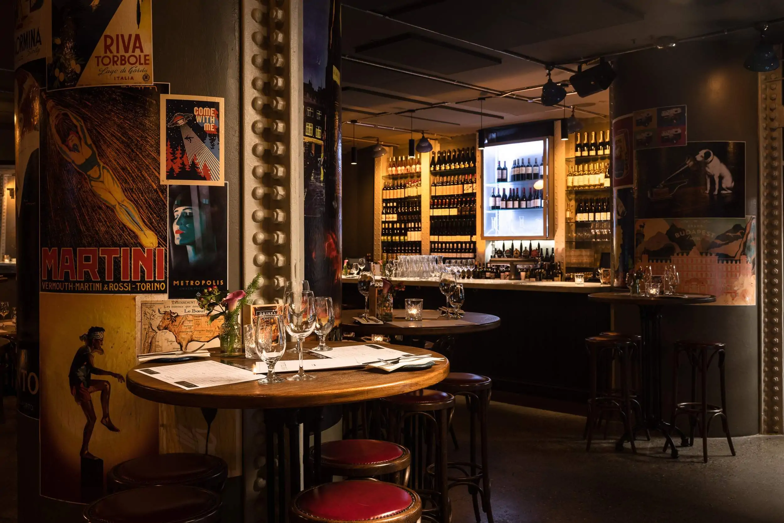 The dimly lit wine cellar at upmarket grocery store Fallon & Byrne in Dublin features high tables with wine glasses, red-topped stools, and wall-to-wall wine bottles. Nouveau French art posters cover the walls.